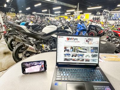 Desk in a motorcycle showroom with a laptop and smartphone ready to film, capturing the perspective of a seller preparing to create a video ad on Vidifye.